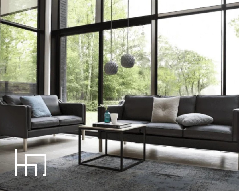 Nowoczesny salon z szarą sofą i fotelami, stolikiem kawowym na tle dużych okien z widokiem na zieleń. Minimalistyczne lampy wiszące dopełniają aranżację.