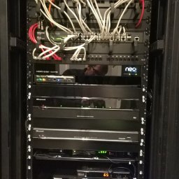 KRC TRADING GROUP SP. Z O.O. - Czarna szafa rack z zainstalowanymi urządzeniami sieciowymi i audio-wideo, widoczne kable, switche, wzmacniacze i inne komponenty. Refleksja osoby w szkle.