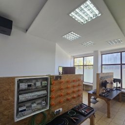 KRC TRADING GROUP SP. Z O.O. - Szafa sterownicza z osprzętem elektrycznym na płycie OSB w jasnym pomieszczeniu z oknami. Widoczne narzędzia i elementy instalacji elektrycznej.