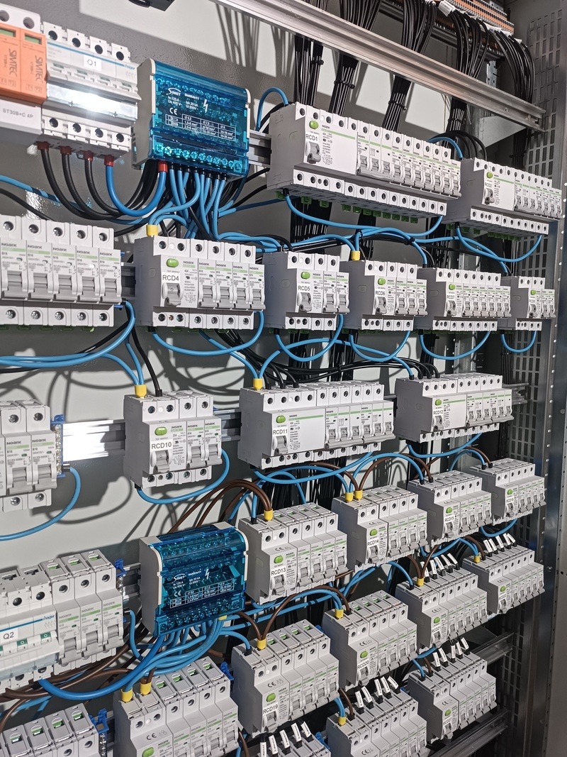 Rozdzielnica elektryczna z licznymi zabezpieczeniami RCD i przewodami w kolorze niebieskim i czarnym, zamontowana w szafie. Widok z bliska na elementy instalacji.