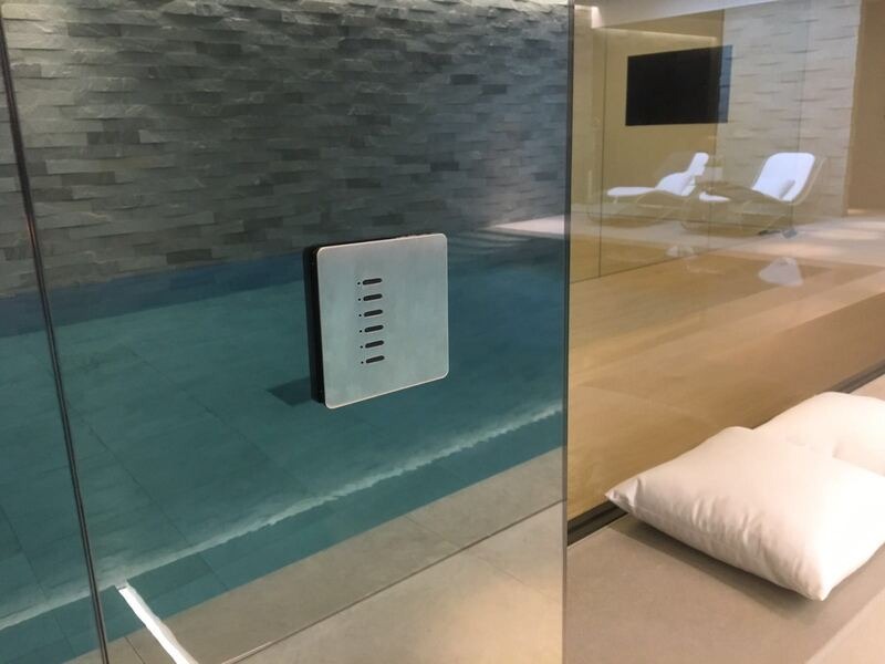 Szklana ściana basenu z panelem sterowania oświetleniem i temperaturą wody. W tle leżaki i telewizor. Nowoczesny design.