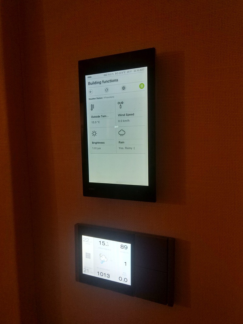 Dwa panele sterowania inteligentnego domu na ścianie: górny z informacjami o pogodzie (temp., wiatr, deszcz), dolny z datą i innymi parametrami. Minimalistyczny design.