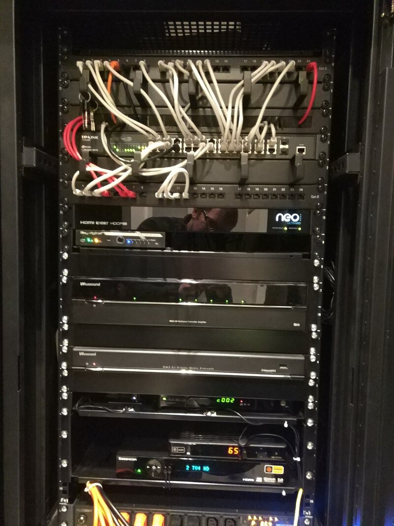 Czarna szafa rack z zainstalowanymi urządzeniami sieciowymi i audio-wideo, widoczne kable, switche, wzmacniacze i inne komponenty. Refleksja osoby w szkle.