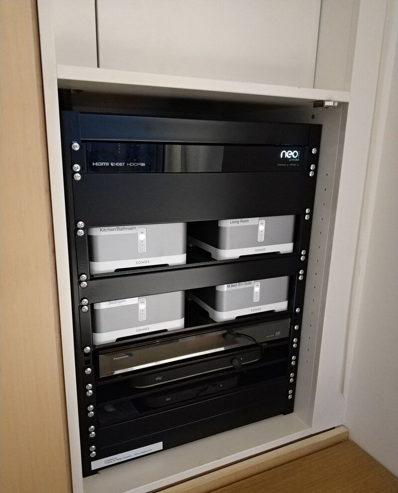 Szafa rack z urządzeniami audio (Sonos) i wideo (Sky, Panasonic), uporządkowana instalacja w zabudowie meblowej, widoczne kable. Integracja systemów audio-wideo.