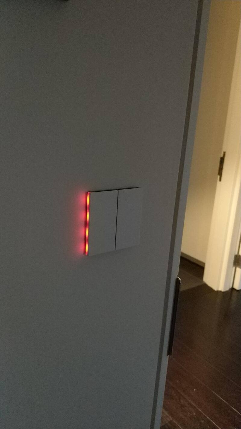 Nowoczesny, podwójny włącznik światła z subtelnym, czerwonym podświetleniem LED, zamontowany na szarej ścianie, widoczny fragment drzwi i podłogi w tle.