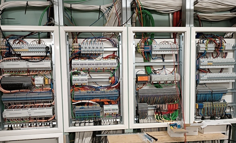 Cztery rozdzielnice elektryczne w białych obudowach, z widocznymi przewodami, bezpiecznikami i elementami sterującymi. Kable ułożone w korytkach. Widok z bliska.