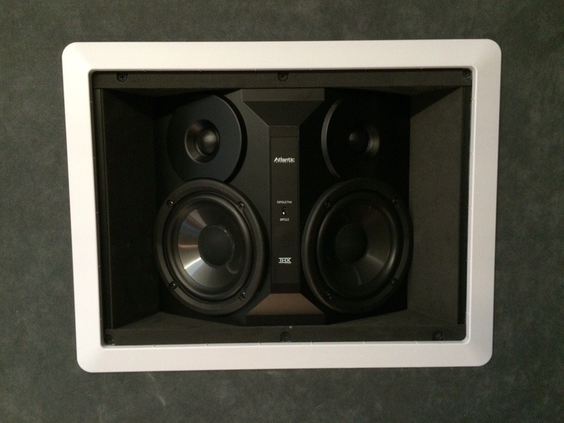 Wbudowany w ścianę głośnik audio marki Atlantic Technology, model IW-260, z białą ramką, widoczny z bliska, na tle szarej ściany. Dwa głośniki niskotonowe i dwa wysokotonowe.