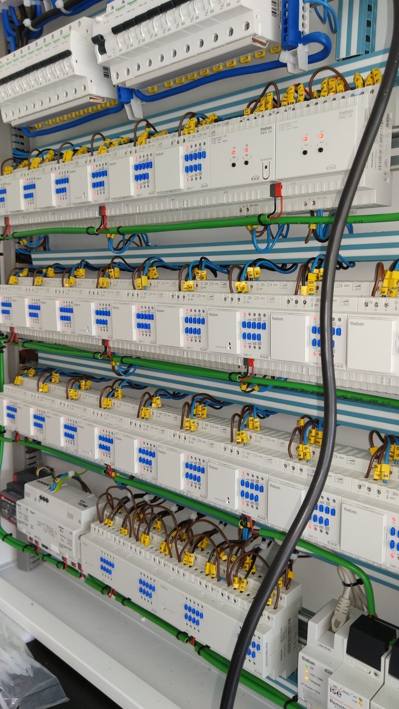 Rozbudowana szafa sterownicza inteligentnego domu z modułami Theben KNX, okablowaniem w peszlach i listwami DIN. Widoczne liczne złącza i diody sygnalizacyjne.