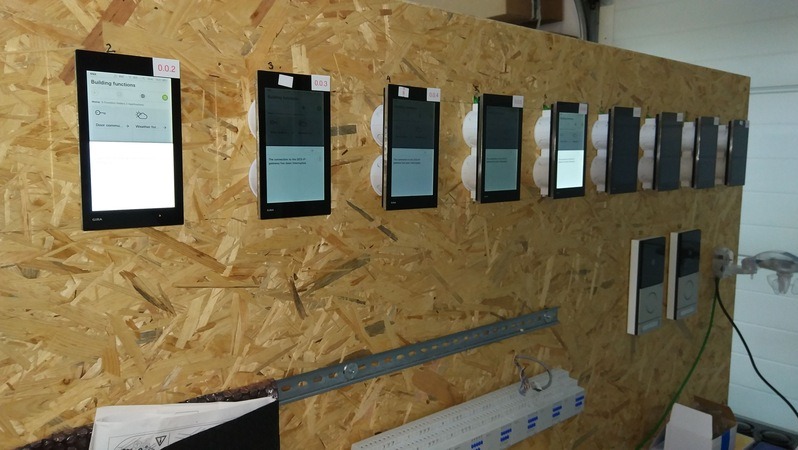 Rząd zamontowanych paneli sterowania inteligentnego domu na płycie OSB, prezentujących interfejs użytkownika i funkcje budynku. Widoczne przewody i elementy montażowe.