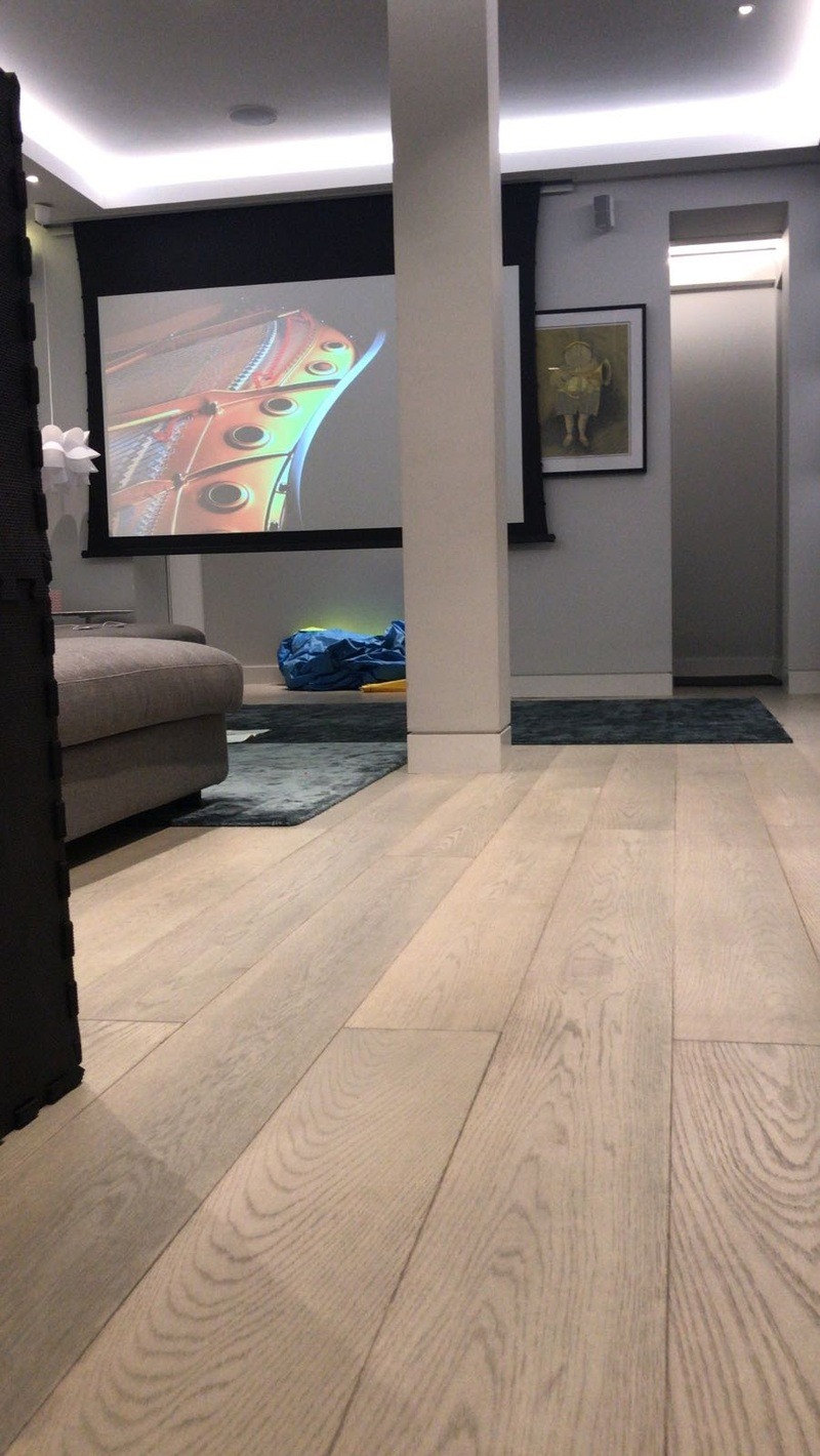 Salon z projektorem i ekranem, widok z dołu na jasną podłogę. Na ścianie obraz w ramie. Częściowo widoczna sofa i kolumna. Subtelne oświetlenie sufitowe.