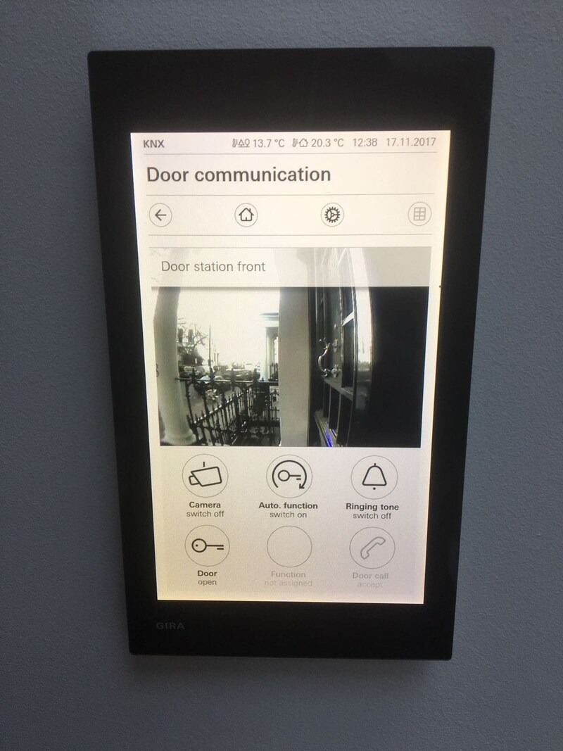 Ekran dotykowy GIRA z interfejsem 'Door communication', prezentujący obraz z kamery przy drzwiach wejściowych i opcje sterowania, takie jak otwieranie drzwi i wyciszanie dzwonka.