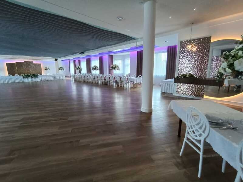 Przestronna sala bankietowa z parkietem, oświetleniem LED i dekoracjami kwiatowymi. Widoczne stoły z białymi obrusami i krzesłami, kolumna oraz dekoracyjne panele ścienne.