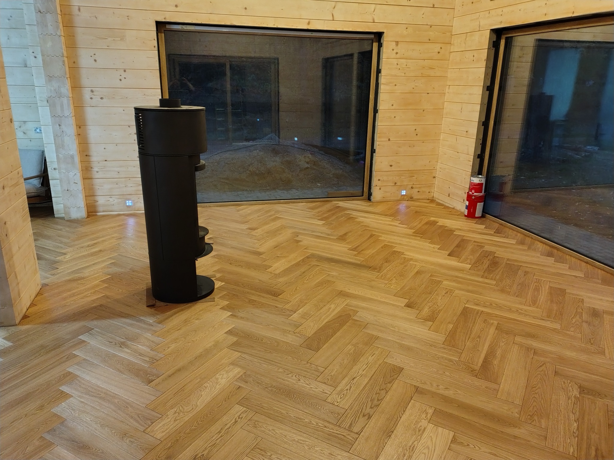 Jasna, drewniana podłoga ułożona w jodełkę francuską w przestronnym wnętrzu z drewnianymi ścianami i dużymi oknami. Minimalistyczny piec wolnostojący.