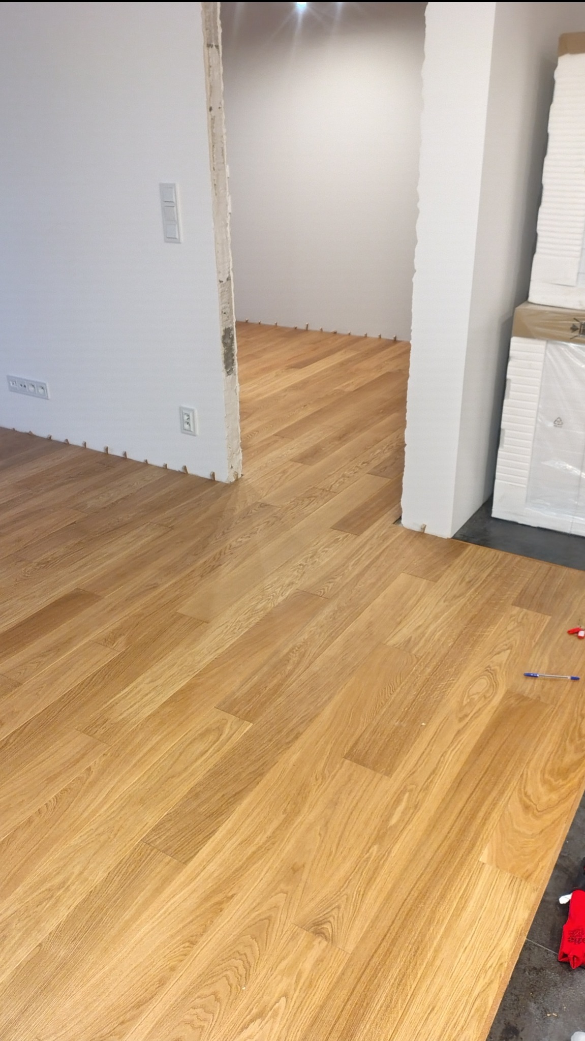 Jasna, drewniana podłoga w trakcie montażu, widoczne kliny dystansowe przy ścianach. Częściowo ułożona podłoga w przejściu do innego pomieszczenia.