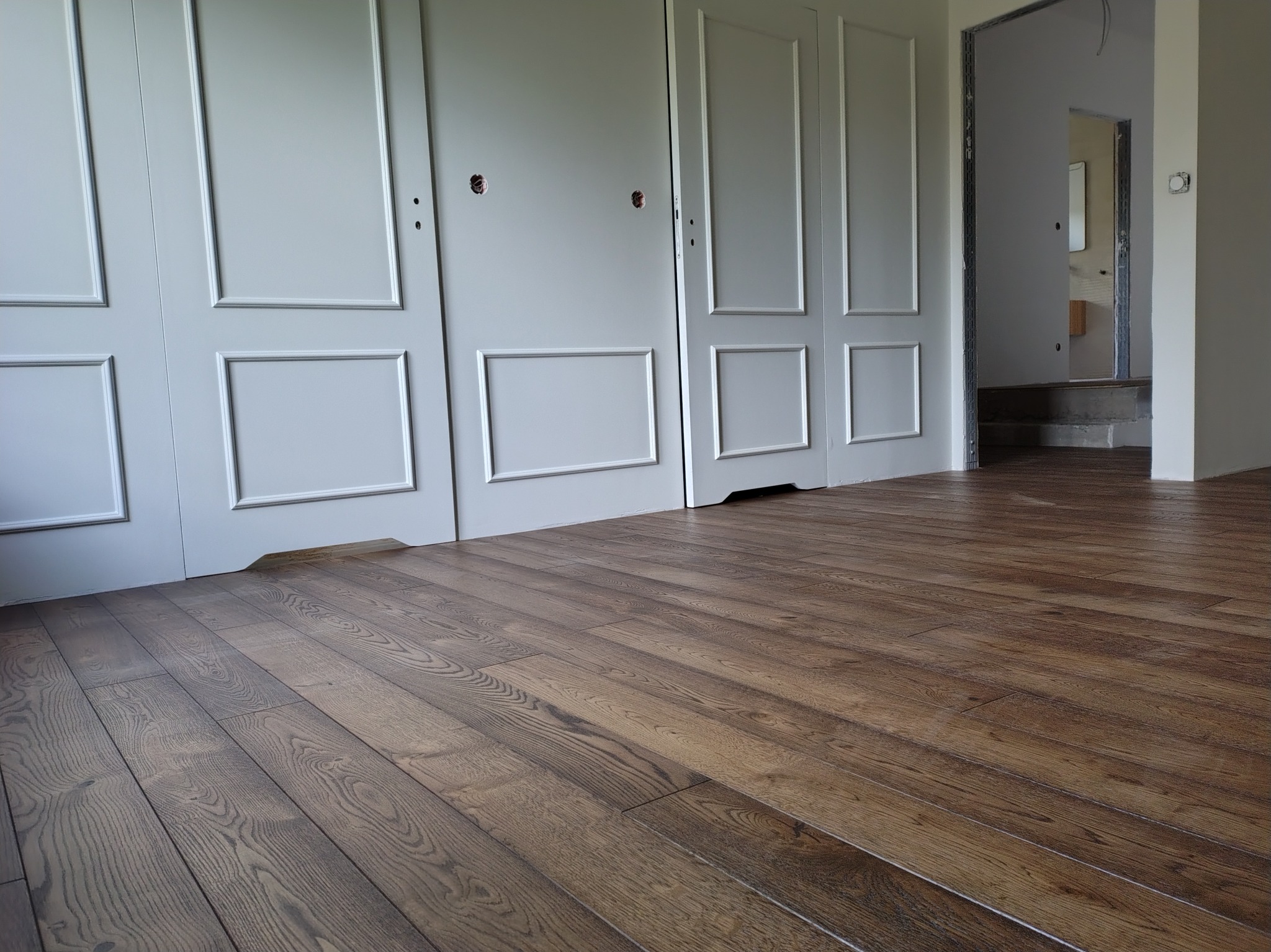 Ukończona, ciemna podłoga drewniana z wyraźnym usłojeniem, ułożona w nowym wnętrzu z białymi, panelowymi drzwiami i widokiem na schody.