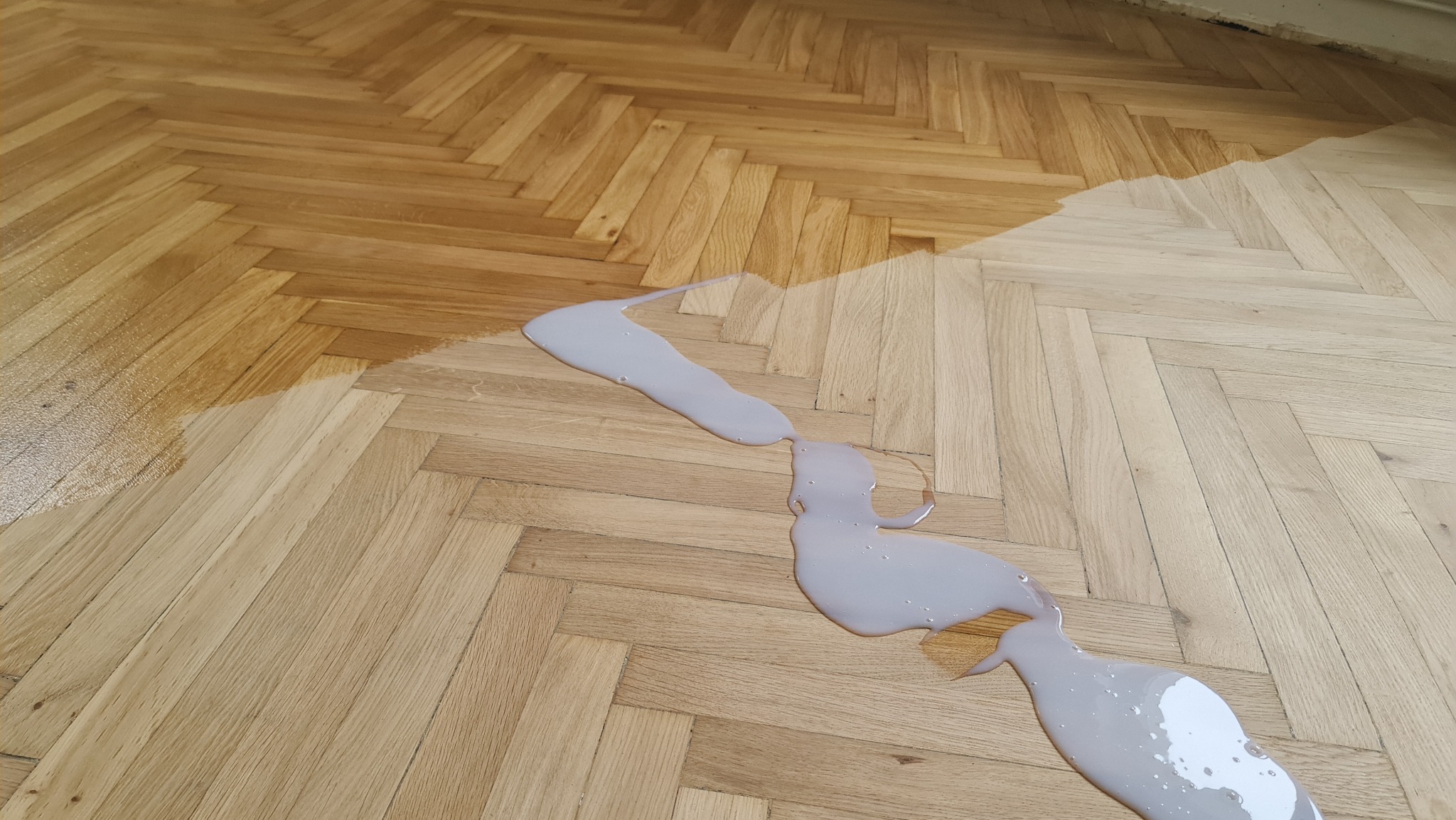 Parkiet w jodełkę, częściowo polakierowany na brązowo, z rozlaną szarą farbą. Widoczny kontrast między surowym a zabezpieczonym drewnem.