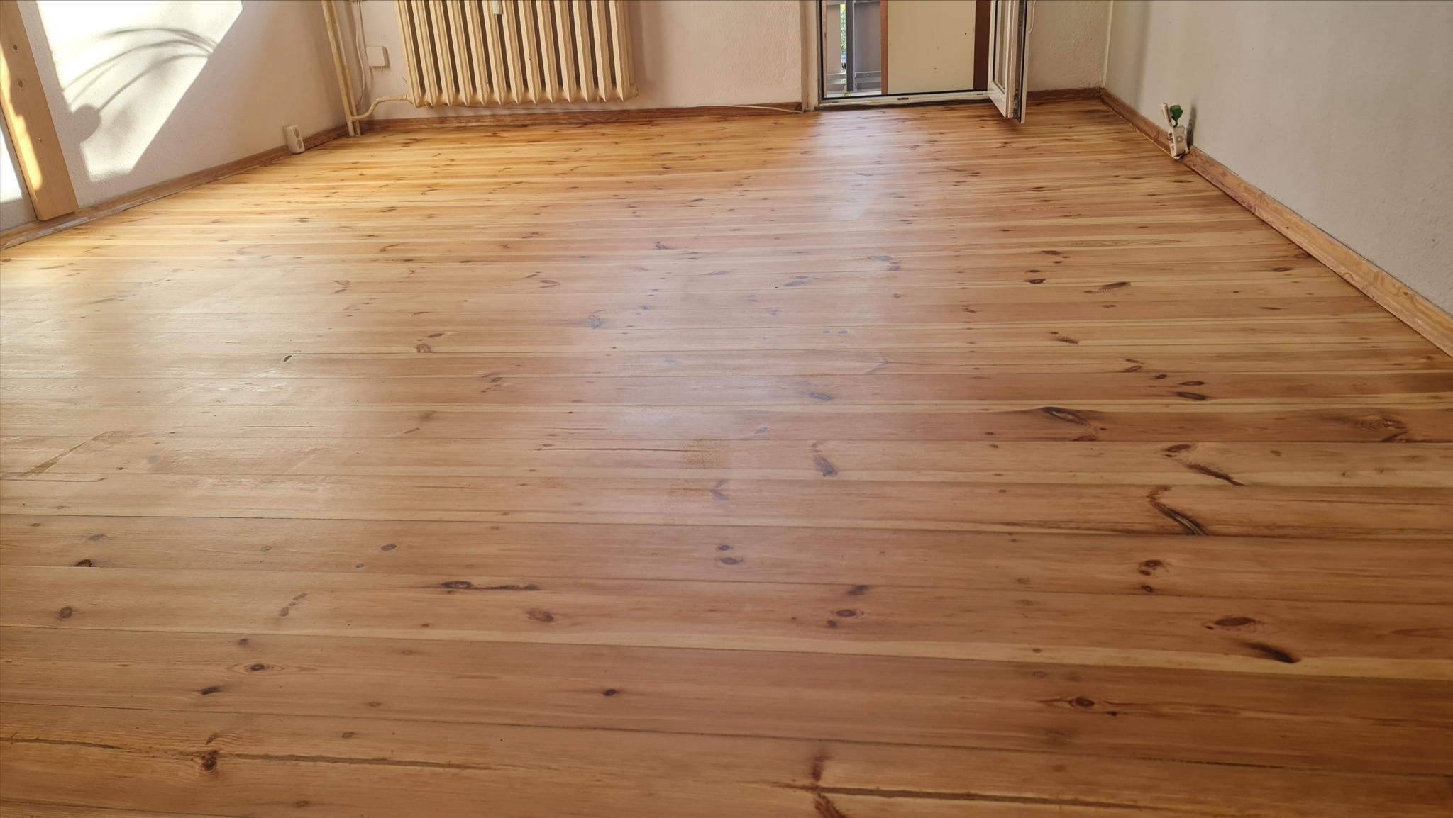 Jasna, lakierowana podłoga z desek sosnowych w pustym pokoju, widok z góry. Naturalne usłojenie drewna, listwy przypodłogowe i grzejnik żeliwny przy ścianie.