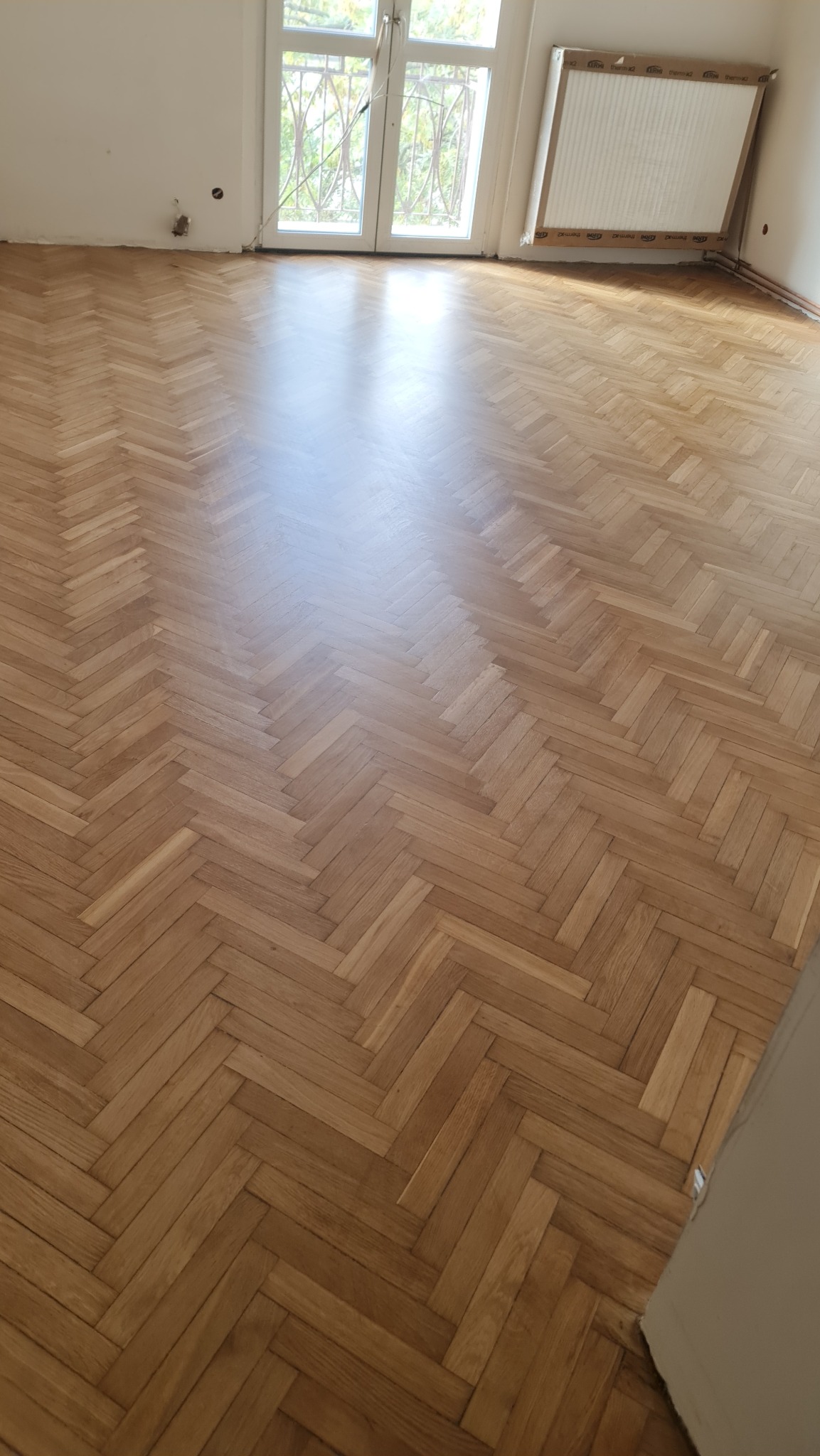 Ułożony parkiet w jodełkę francuską, widok na jasne, przestronne wnętrze z oknem balkonowym i grzejnikiem. Parkiet świeżo po lakierowaniu, z połyskiem.