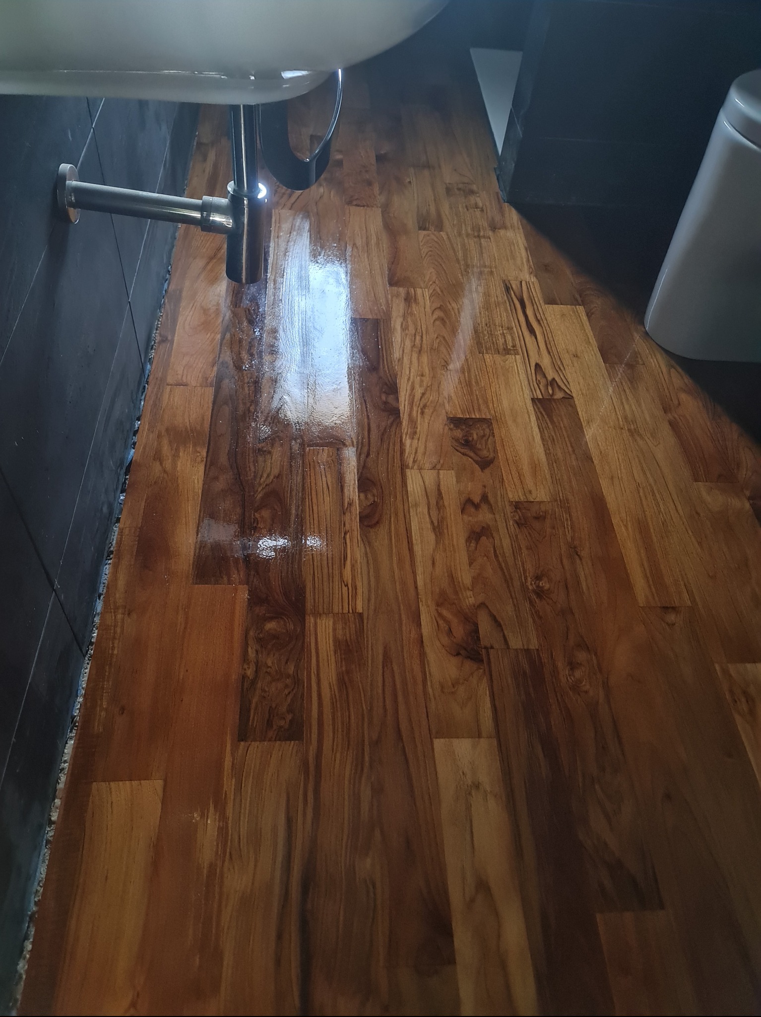 Ułożone panele w łazience z widoczną instalacją umywalki i fragmentem toalety. Brązowe panele z wyraźnym wzorem drewna, szara ściana i biała ceramika.