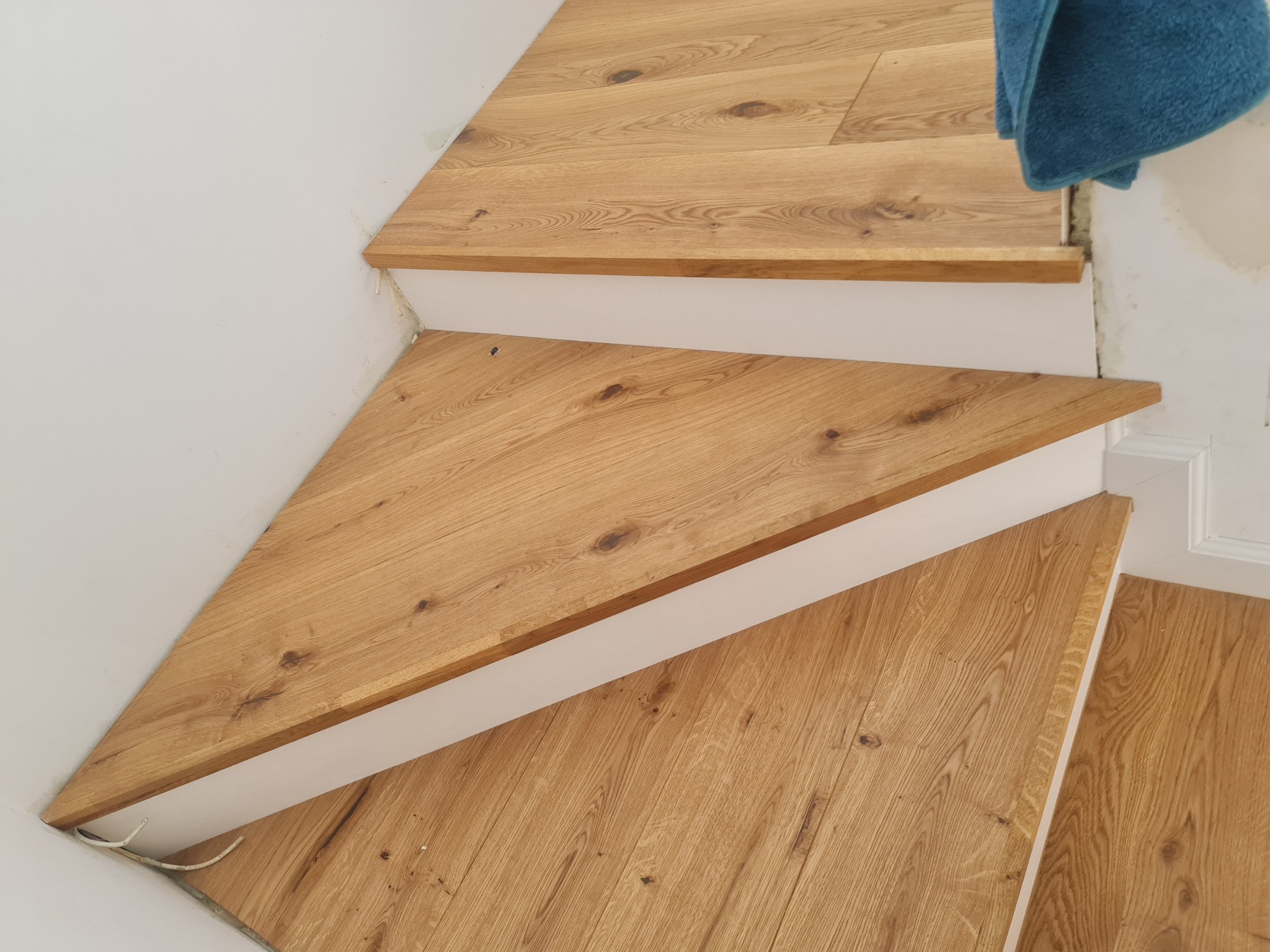 Wykończone drewniane schody z jasnego drewna dębowego, widok z góry, z fragmentem niebieskiego materiału w tle. Detal wykończenia wnętrza.