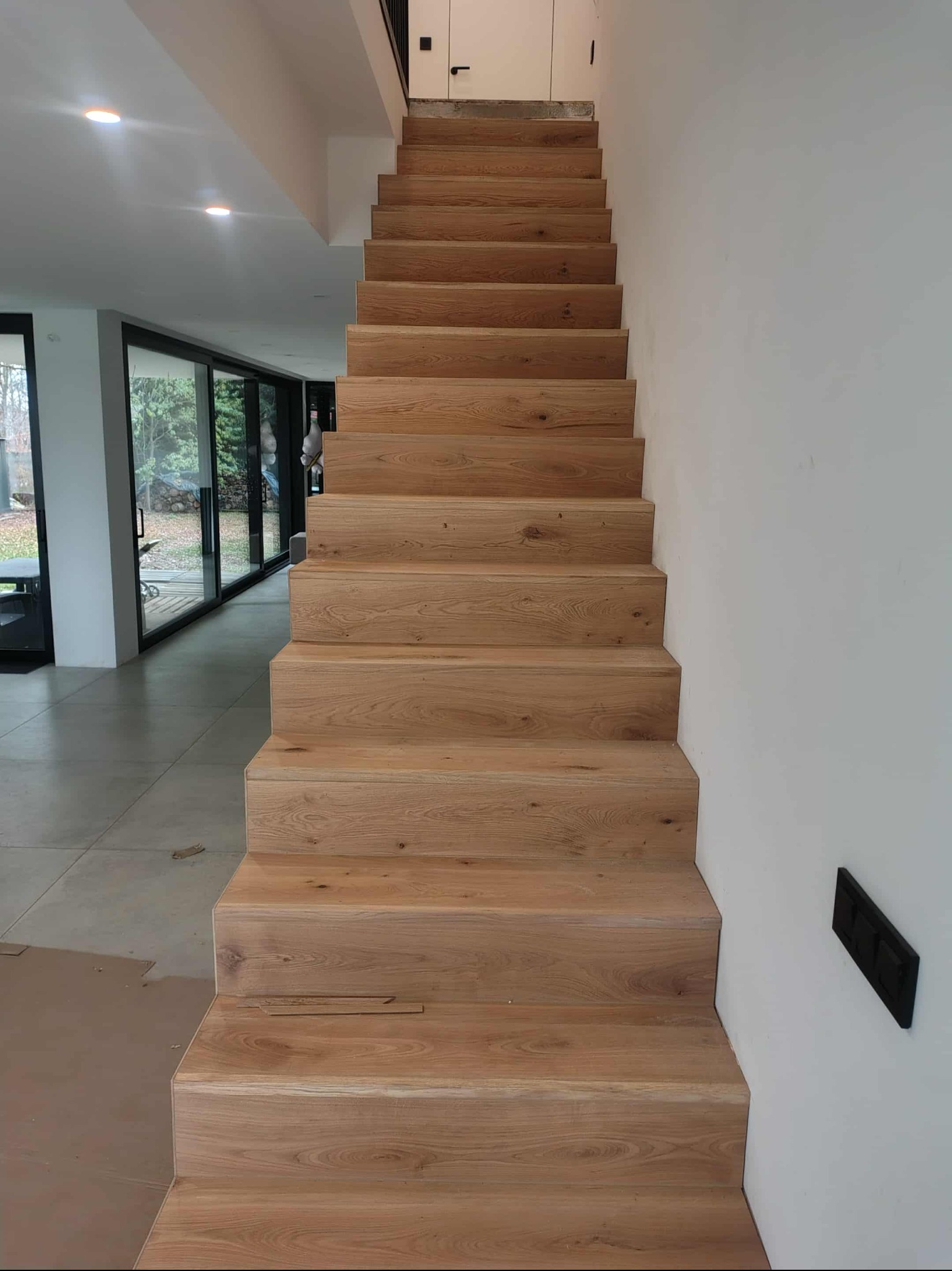 Drewniane schody z jasnego drewna, prowadzące do góry, z widoczną strukturą słojów. Minimalistyczne wnętrze z dużymi oknami i białymi ścianami. Nowoczesny design.