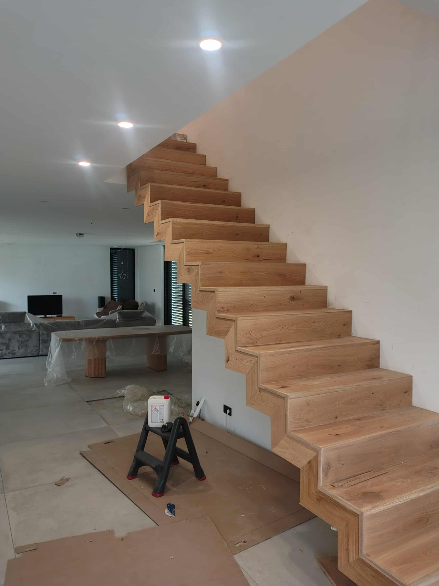 Montaż drewnianych schodów w nowoczesnym wnętrzu. Widoczne stopnie z jasnego drewna, drabina malarska i zabezpieczone meble. Oświetlenie punktowe na suficie.
