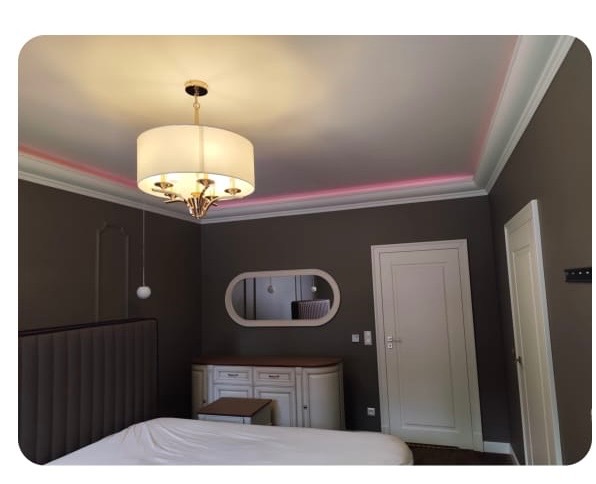 Pokój z szarą ścianą, białymi drzwiami i listwami, oświetleniem LED pod sufitem i lampą wiszącą. Widoczne łóżko i komoda z lustrem.