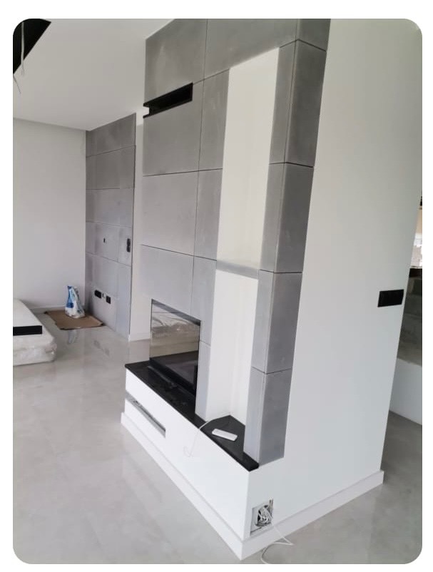 Nowoczesny kominek obudowany szarymi płytami i białymi elementami, z czarnym blatem. Widoczne gniazdka elektryczne i fragment podłogi z połyskiem.