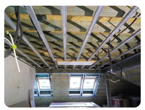 Montaż sufitu podwieszanego na poddaszu z oknami dachowymi. Widoczne metalowe profile, izolacja i instalacje. Etap prac przed zamknięciem płytami gipsowo-kartonowymi.