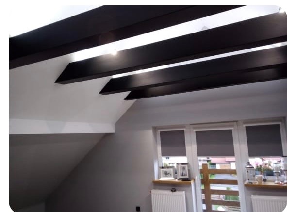 Nowoczesny sufit z ciemnymi belkami w jasnym wnętrzu. Widoczne okna z roletami i skos dachu. Minimalistyczny design.