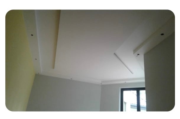 Biały sufit podwieszany z dekoracyjnymi listwami w nowocześnie wykończonym wnętrzu. Widoczne punkty świetlne i fragment ściany w odcieniach szarości i żółci.