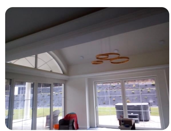 Jasne wnętrze z dużymi oknami i nowoczesnym oświetleniem. Widoczne białe ściany, sufit z belkami i okrągła lampa. Na zewnątrz zielony trawnik i elementy architektury ogrodowej.