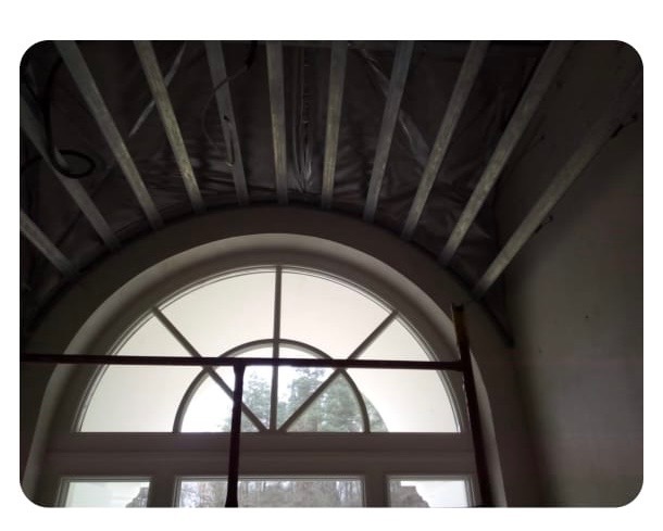 Montaż sufitu podwieszanego z metalową konstrukcją nad łukowym oknem. Widoczne elementy instalacji i rusztowanie.