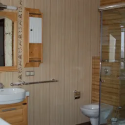 Jasna łazienka z drewnianymi akcentami: szafka pod umywalką, lustro z szafką, kabina prysznicowa ze szklanymi drzwiami, toaleta i beżowe płytki na ścianach.