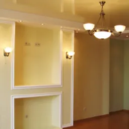 Jasne wnętrze pokoju z wnękami obudowanymi ozdobnymi listwami, oświetleniem punktowym i klasycznym żyrandolem, ukazujące efekt prac wykończeniowych.