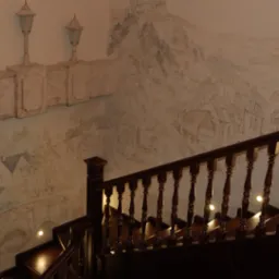 Klatka schodowa z ciemnego drewna z balustradą z toczonymi tralkami, oświetlona punktowymi lampkami schodowymi, na ścianie dekoracyjna płaskorzeźba i malowidło przedstawiające pejzaż miejski...