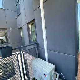 THINK OZE PATRYK SOBOŃ - Zamontowana jednostka zewnętrzna klimatyzacji Gree na balkonie nowoczesnego budynku. Widoczne rury odprowadzające. Sztuczna trawa na podłodze balkonu.