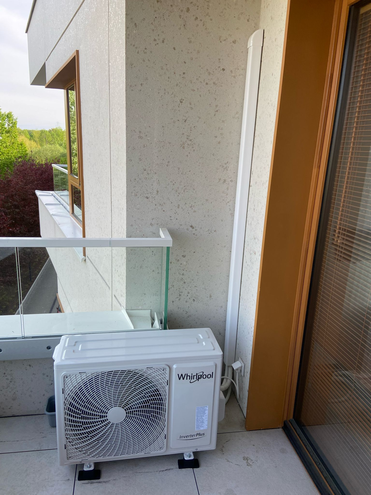 Biała jednostka zewnętrzna klimatyzacji Whirlpool Inverter Plus na balkonie z szklaną balustradą, widok z boku, z elementami elewacji i oknem w tle.
