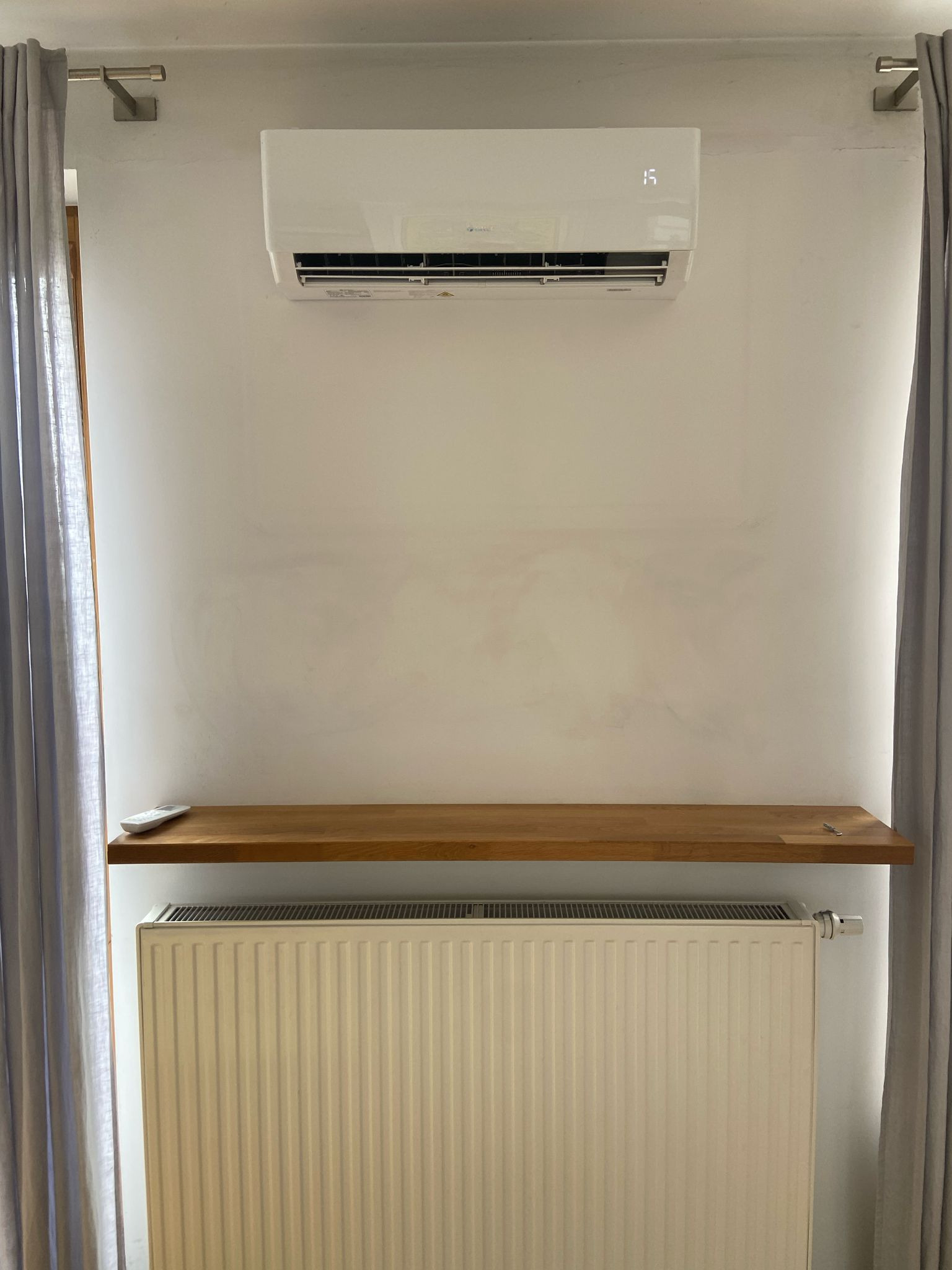 Biały klimatyzator ścienny z wyświetlaczem temperatury, zamontowany nad grzejnikiem i drewnianą półką. Zasłony po bokach kadru.