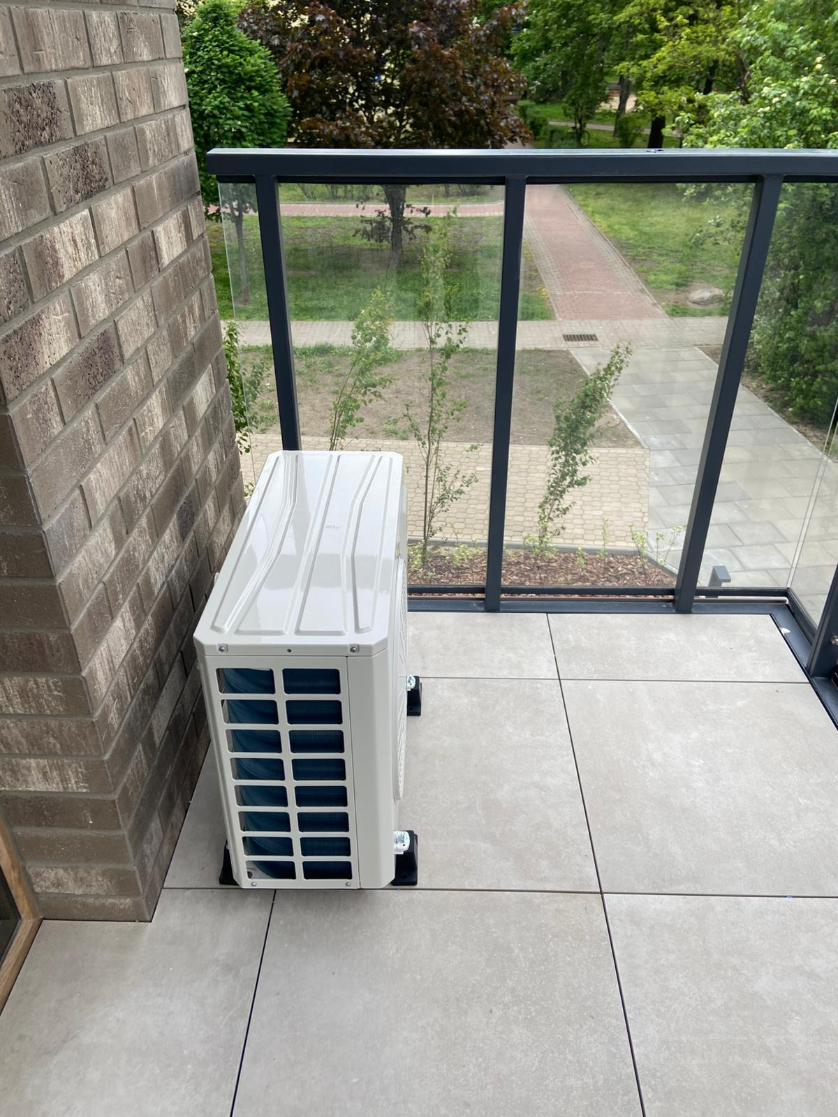 Biała jednostka zewnętrzna klimatyzacji na balkonie z szarą podłogą i szklaną balustradą, widok na zieleń. Montaż klimatyzacji na zewnątrz budynku.