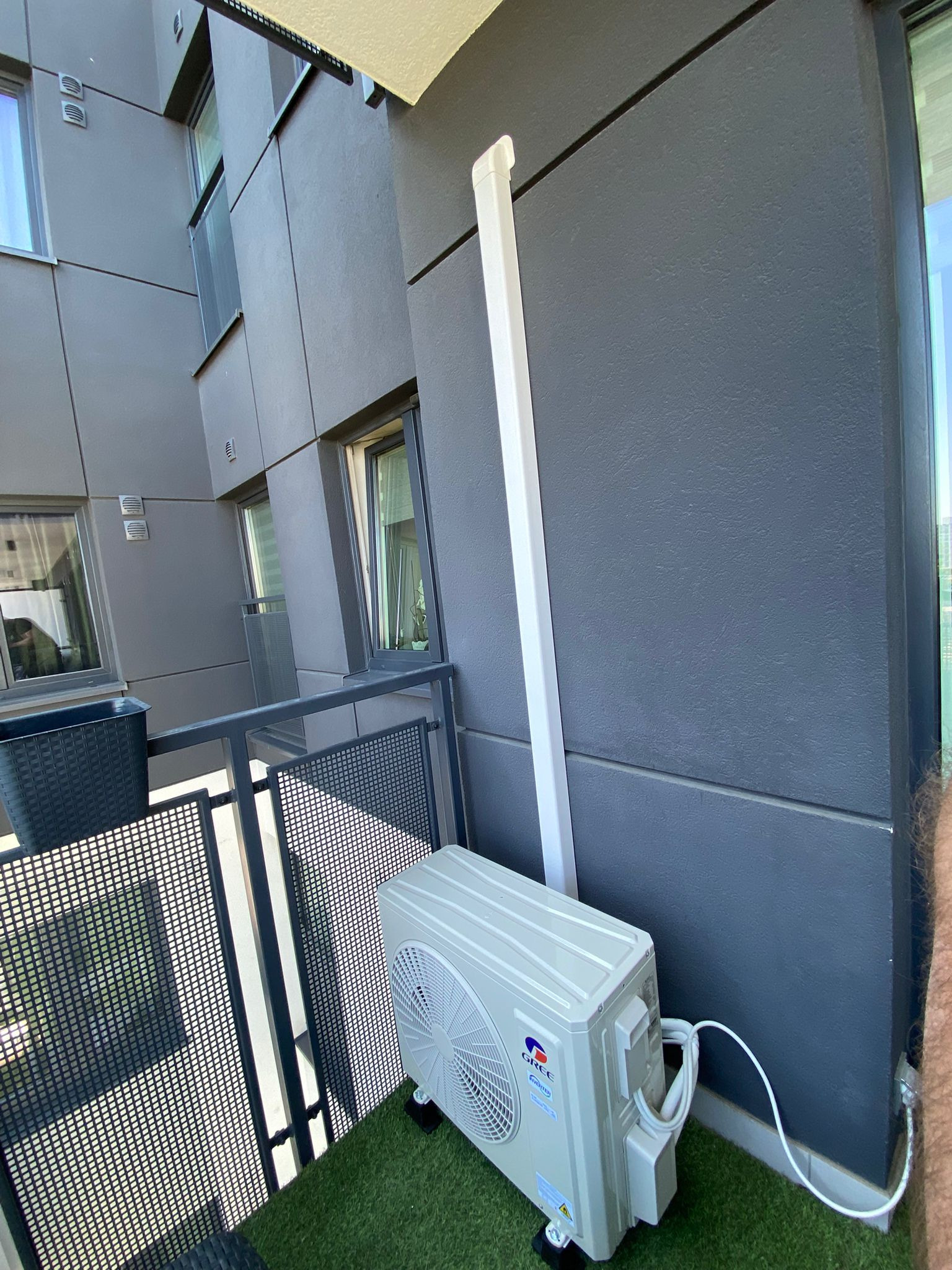 Zamontowana jednostka zewnętrzna klimatyzacji Gree na balkonie nowoczesnego budynku. Widoczne rury odprowadzające. Sztuczna trawa na podłodze balkonu.
