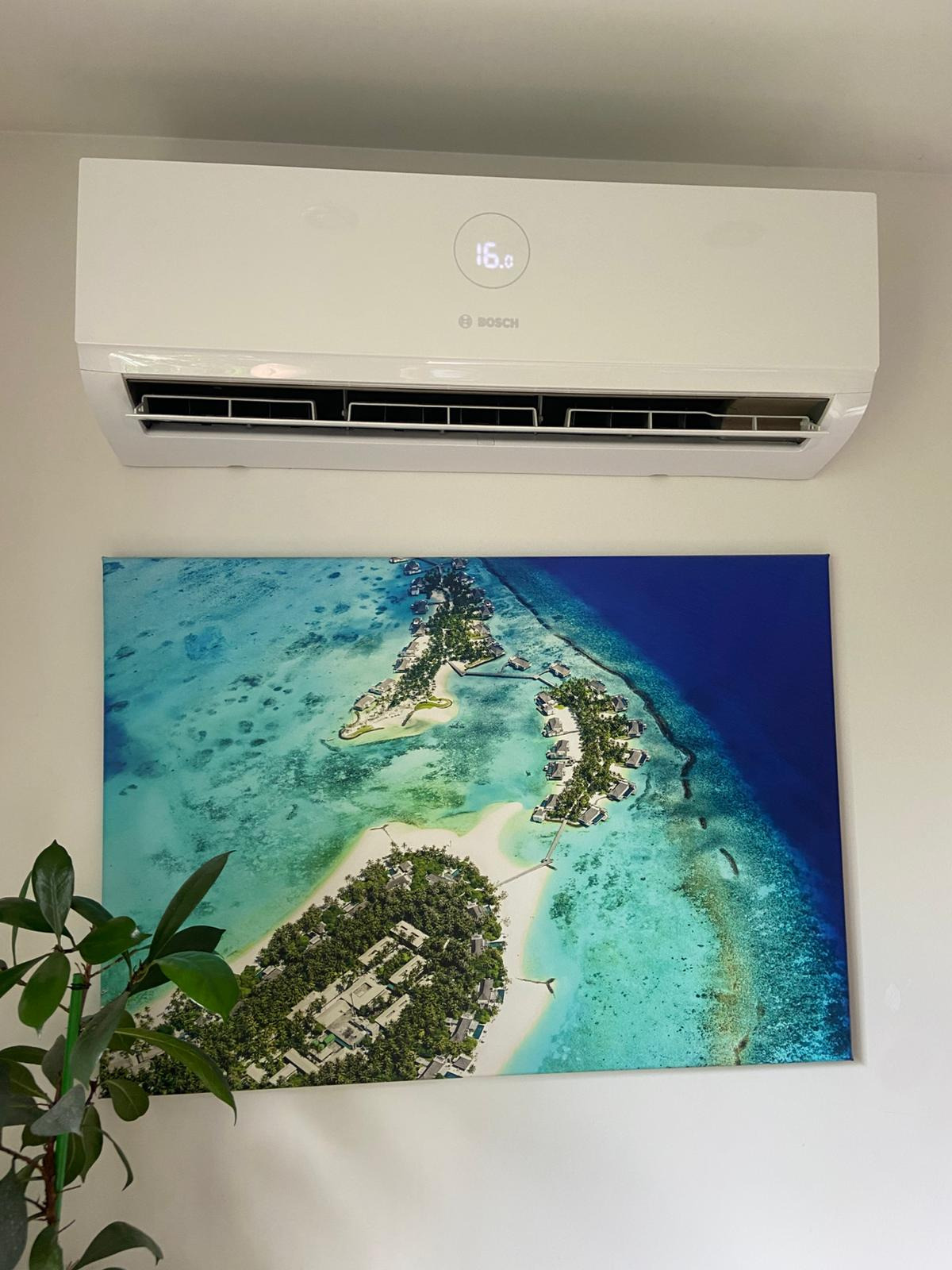 Biały klimatyzator Bosch na ścianie nad obrazem z tropikalną wyspą. Widoczny fragment rośliny doniczkowej po lewej stronie.