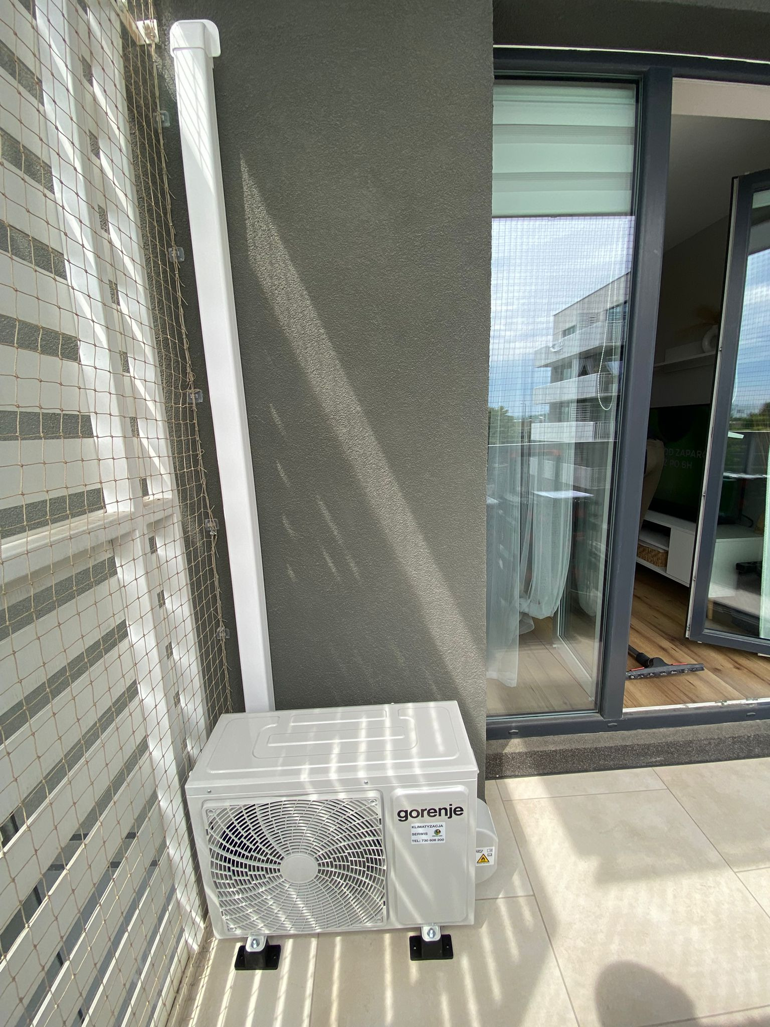 Biała jednostka zewnętrzna klimatyzacji Gorenje zamontowana na balkonie, widoczna krata balkonu i fragment otwartych drzwi balkonowych.