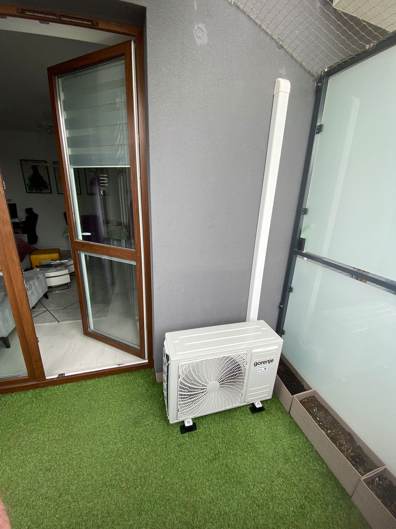 Biała jednostka zewnętrzna klimatyzacji Gorenje na balkonie z sztuczną trawą, obok okna balkonowego i donic. Szara ściana w tle.