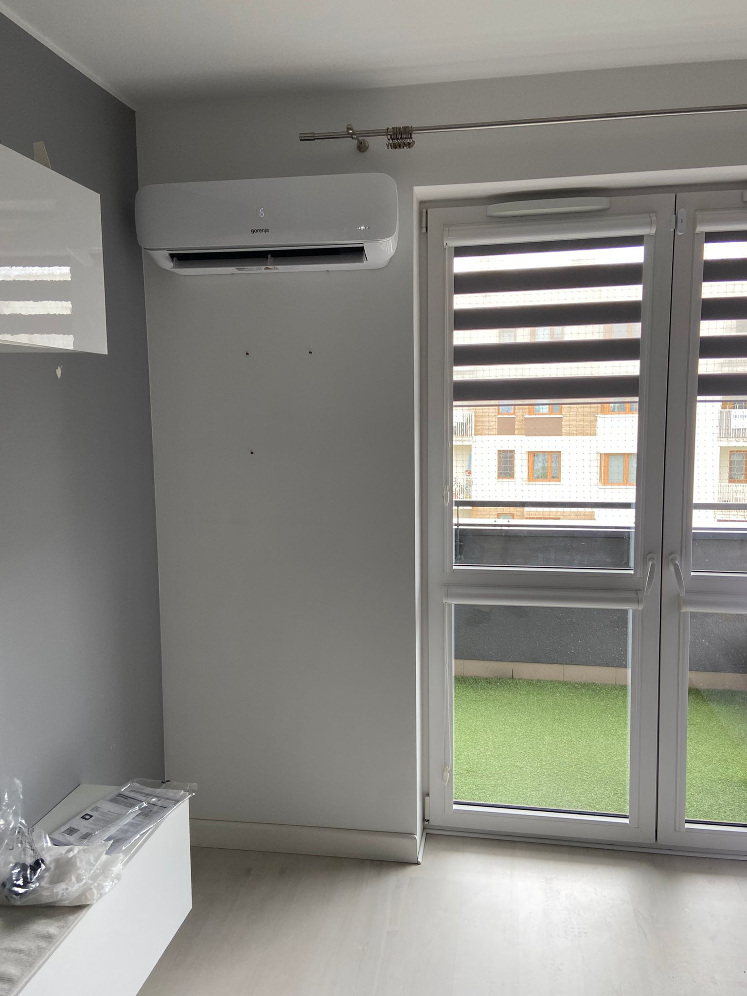 Biały klimatyzator Gorenje zamontowany na ścianie w jasnym pokoju z dużym oknem balkonowym i roletami dzień-noc. Widoczne ślady po montażu.