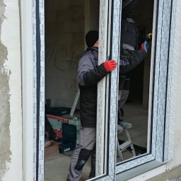 Vision House Jakub Stosio - Montaż okna przesuwnego Schüco przez dwóch monterów na budowie. Widoczne narzędzia i elementy konstrukcyjne. Okno z białymi profilami i czarnymi uszczelkami.