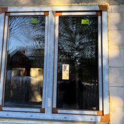 Vision House Jakub Stosio - Nowe, dwuskrzydłowe okno z brązową ramą, zamontowane w surowej ścianie z betonu komórkowego. Widoczne pianka montażowa i folia ochronna na profilach.