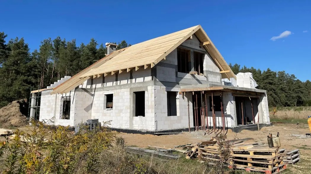 Surowy stan domu z białego bloczka, z częściowo wykonanym drewnianym dachem, na tle lasu i błękitnego nieba. Widoczne rusztowania i materiały budowlane.
