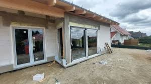 Nowoczesny dom w trakcie budowy z zamontowanymi oknami PCV i roletami zewnętrznymi w kolorze szarym. Widoczne rusztowania i narzędzia budowlane.