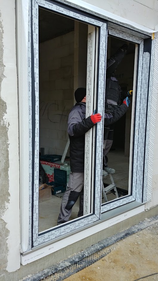 Montaż okna przesuwnego Schüco przez dwóch monterów na budowie. Widoczne narzędzia i elementy konstrukcyjne. Okno z białymi profilami i czarnymi uszczelkami.